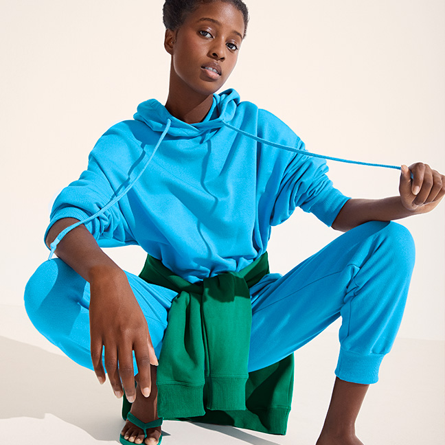Mulher preta abaixada, olhando para a câmera, vestindo um conjunto de moletom hering azul com uma blusa verde amarrada na cintura e uma havaianas verde
