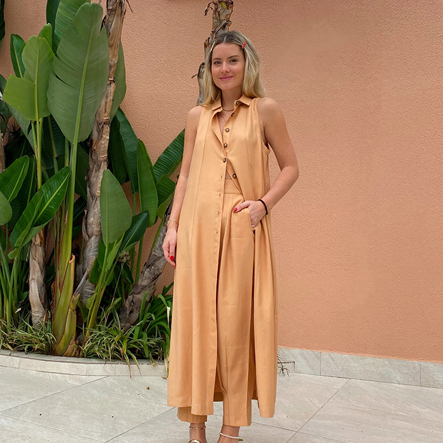 Influenciadora Júlia Tibério em pé , sorrindo para a câmera com a mão no bolso da calça . Ela veste um vestido chemise aberto e uma calça slouchy